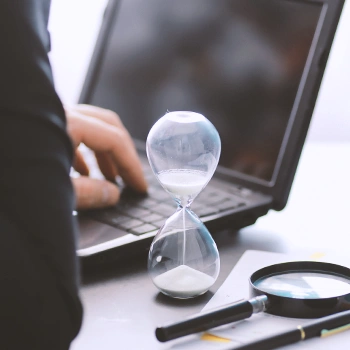 An office table with an hourglass, a laptop, and a magnifying glass