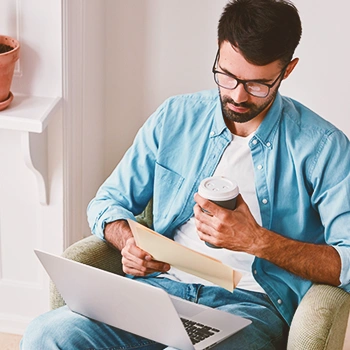 A man holding a piece of paper in front of his laptop while drinking coffee