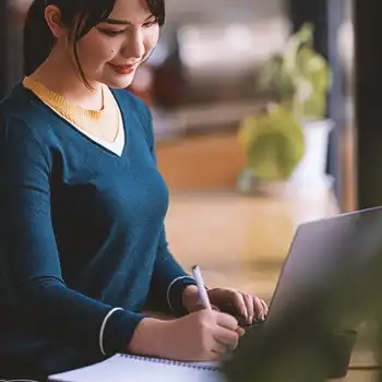 A person writing down notes while in front of a laptop