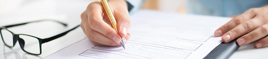 A person writing on a table with eyeglasses on the side