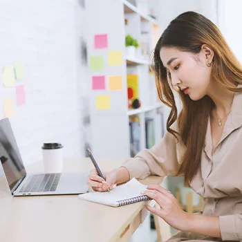 An office worker writing down notes in front of a laptop